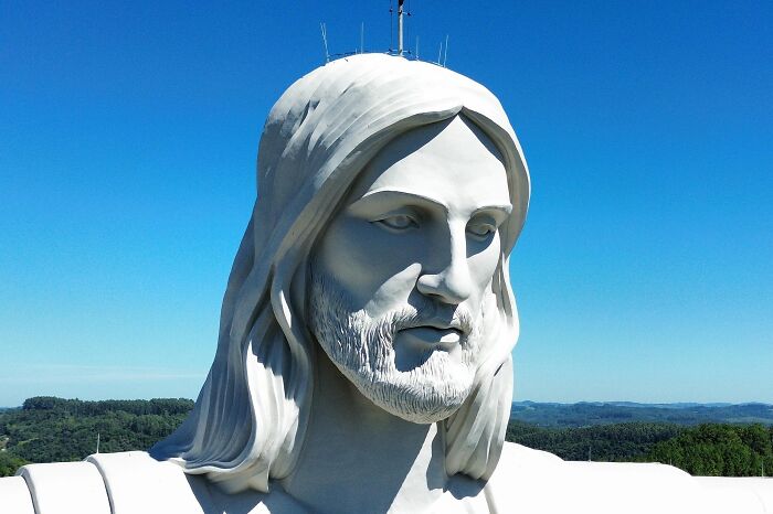 Large white sculpture of a serene bearded man with long hair against a clear blue sky and distant landscape background.