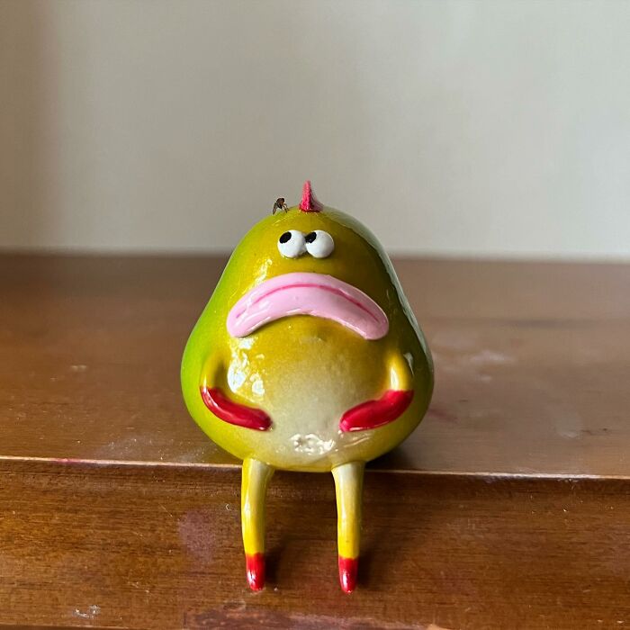 Quirky sculpture of a yellow-green character with pink mouth and red hands sitting on a wooden surface bursting with personality.
