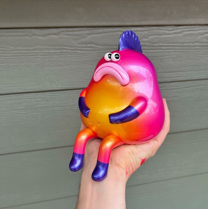 Hand holding a colorful quirky sculpture with expressive eyes and fins, showcasing artist made quirky sculptures bursting with personality.