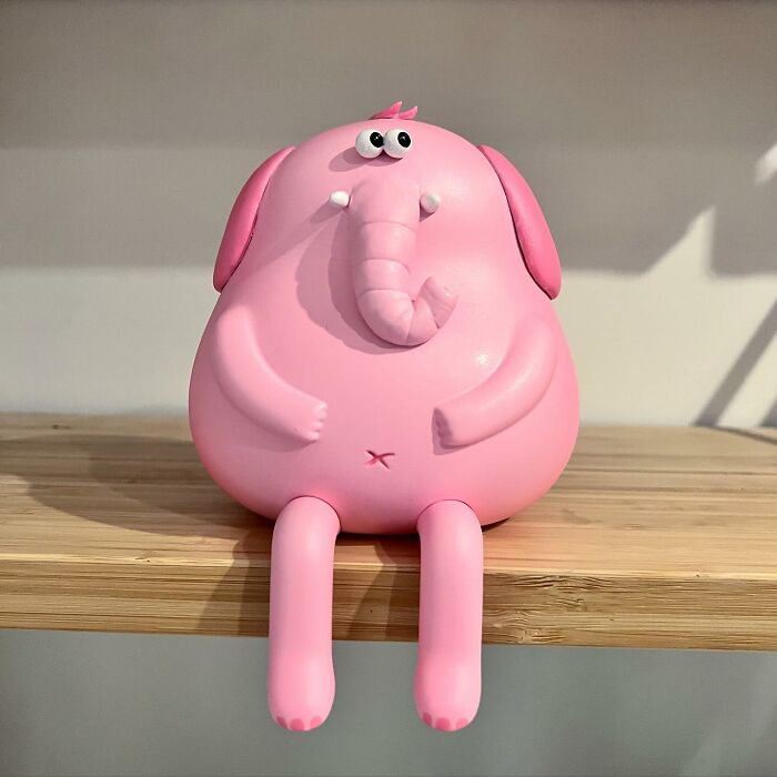 Pink quirky sculpture of an elephant with expressive eyes and rounded body sitting on a wooden surface, bursting with personality.