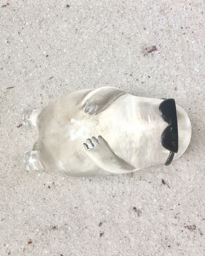 Sculpture of a quirky character lying on sand wearing black sunglasses, showcasing playful personality and artistic design.