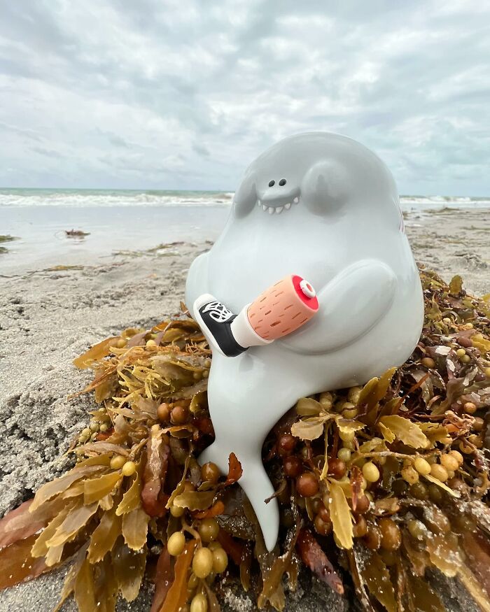 Quirky sculpture of a smiling ghost-like figure holding a sneaker on a seaweed-covered beach by the ocean.