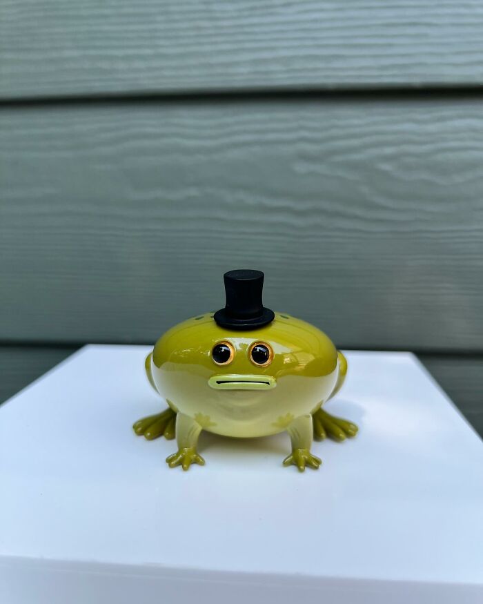 Glossy quirky sculpture of a frog wearing a black top hat bursting with personality on a white surface outdoors.