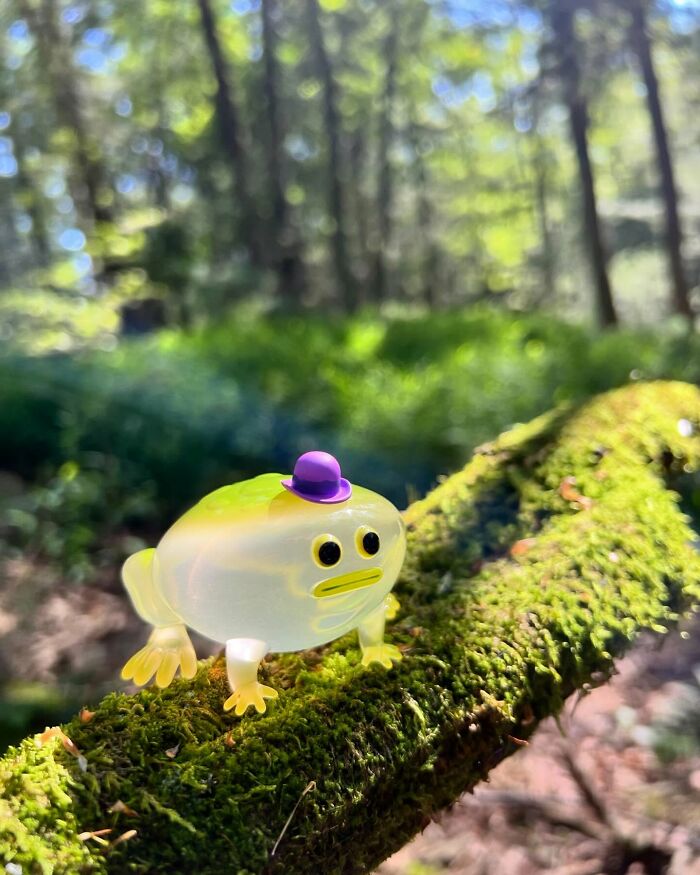 Quirky sculpture of a translucent creature with a purple hat on mossy tree branch in a sunlit forest setting.
