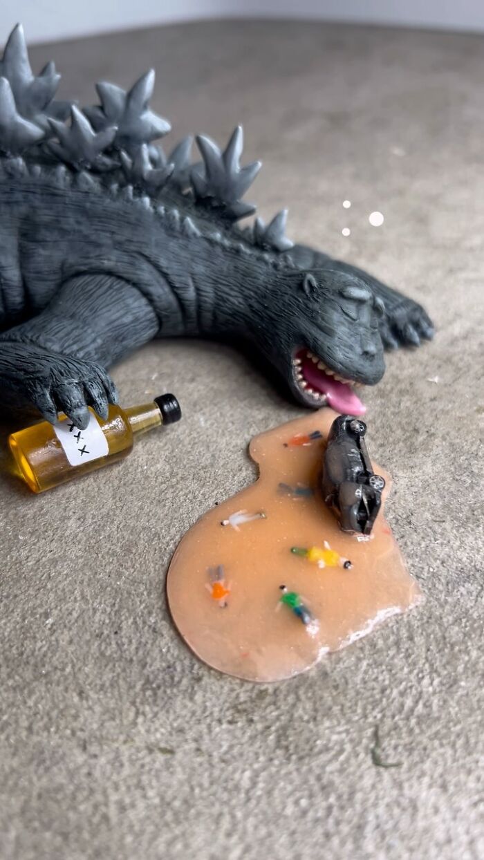 Quirky sculpture of a dragon holding a bottle next to a spilled pool with tiny people and an overturned car.