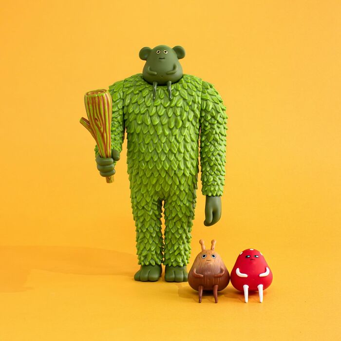 Green quirky sculpture holding a wooden club standing next to two small quirky sculptures against a yellow background.