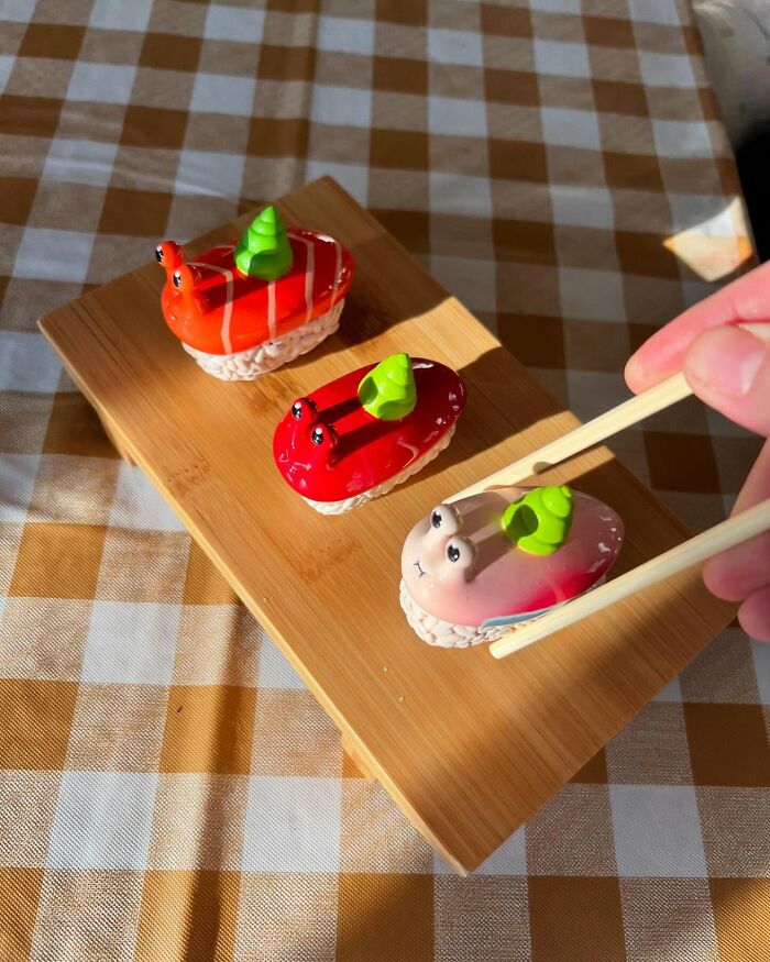 Three quirky sculptures resembling sushi with snail eyes on a wooden board, with chopsticks holding one sculpture.