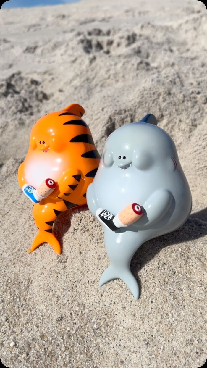 Two quirky sculptures bursting with personality, one orange with black stripes and one gray, sitting on sand holding small objects.