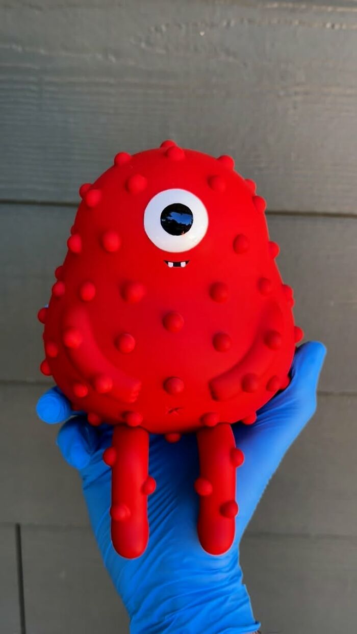 Red quirky sculpture with one eye and textured bumps held by hand in blue glove, showcasing unique artist made personality.