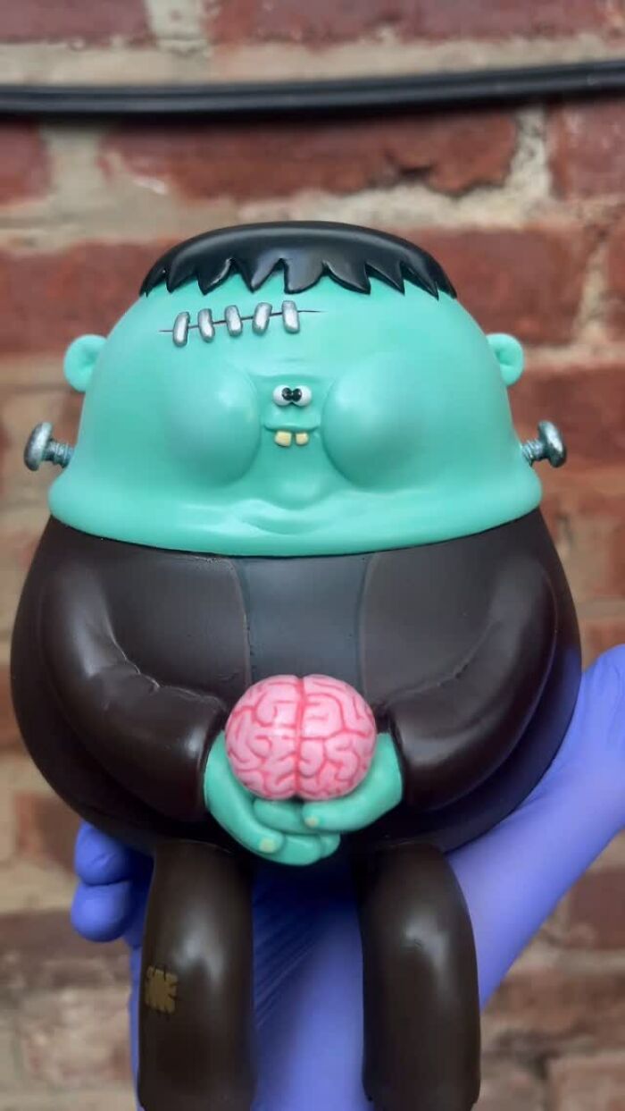 Hand holding quirky sculpture of a green Frankenstein character with bolts and holding a pink brain, bursting with personality.