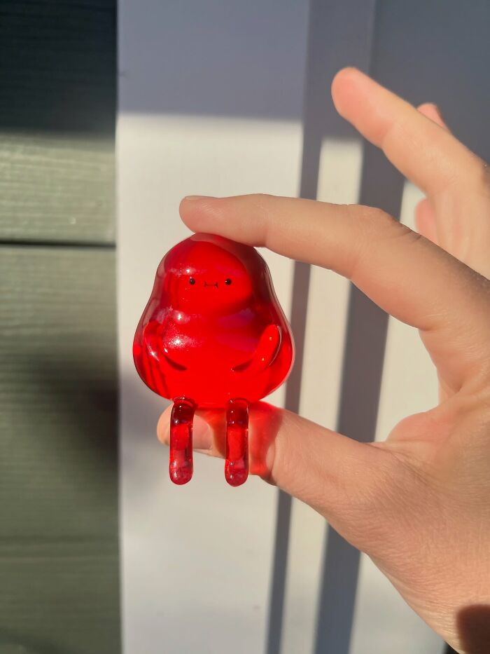 Hand holding a small red quirky sculpture with a cute face and legs, showcasing unique personality in bright sunlight.