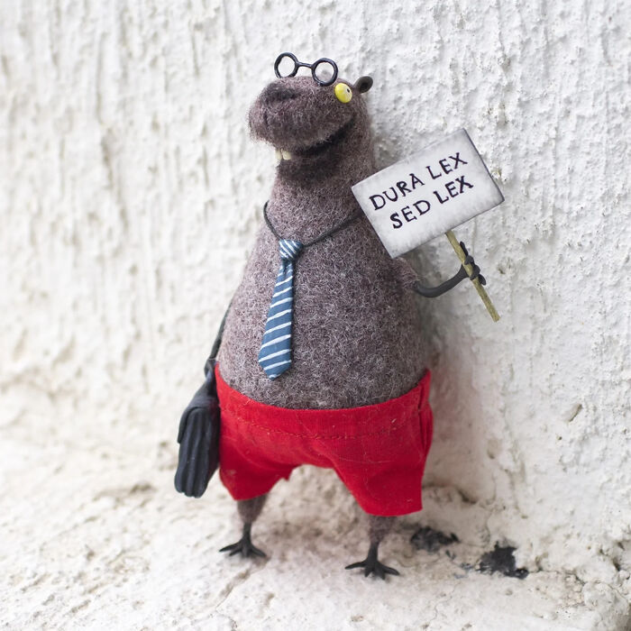 Handmade creature toy with glasses, striped tie, red shorts, holding a sign, standing against a textured wall.