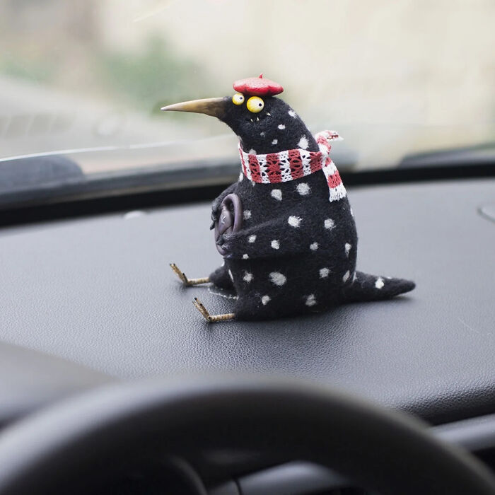 Handmade creature toy resembling a spotted bird wearing a red scarf and beret, sitting on a car dashboard.
