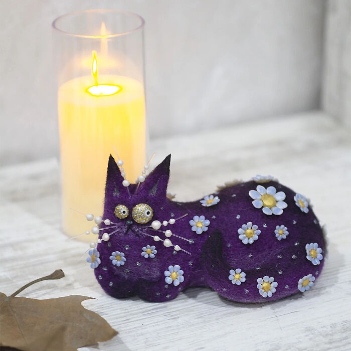 Handmade creature toy shaped like a purple cat decorated with small flowers, displayed near a lit candle on a wooden surface.