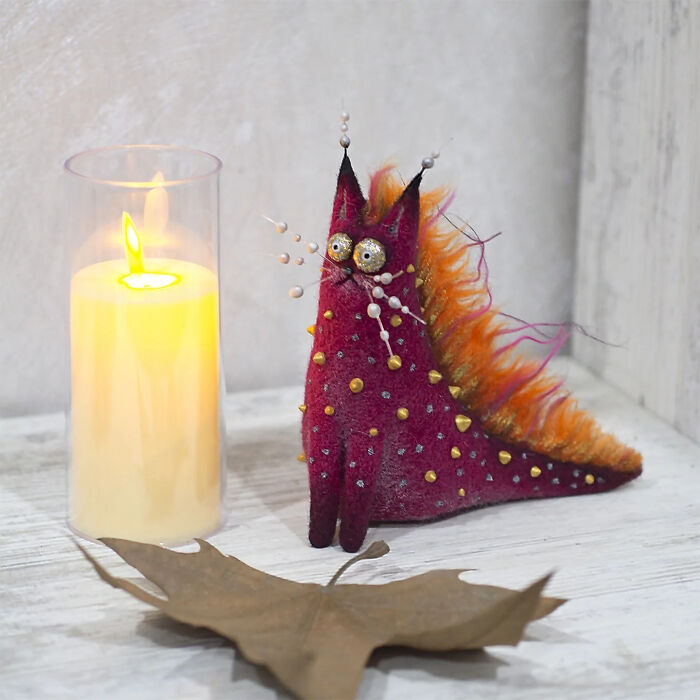 Handmade creature toy with bright orange and purple colors, sitting next to a lit candle and dried leaf on a wooden surface.