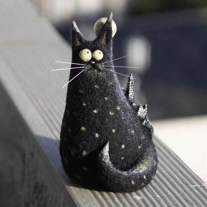 Handmade creature toy resembling a black cat with large eyes and tiny buildings on its back, showcasing adorable strange art.