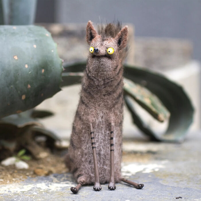 Handmade adorable strange creature toy with yellow eyes sitting on a stone surface near a cactus plant.