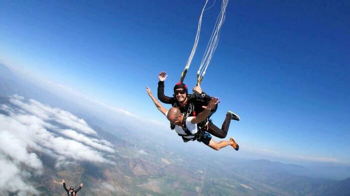 Two men skydiving with one playfully photobombing from below against a clear blue sky and landscape below.