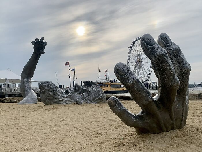 Large hand and face sculptures emerging from sand on a beach near a ferris wheel, showcasing spectacular sculptures globally.