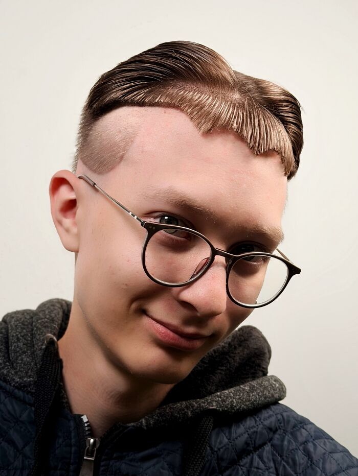 Young man with bold barber haircut featuring sharp lines and artistic styling wearing glasses and a dark hoodie against a white background.