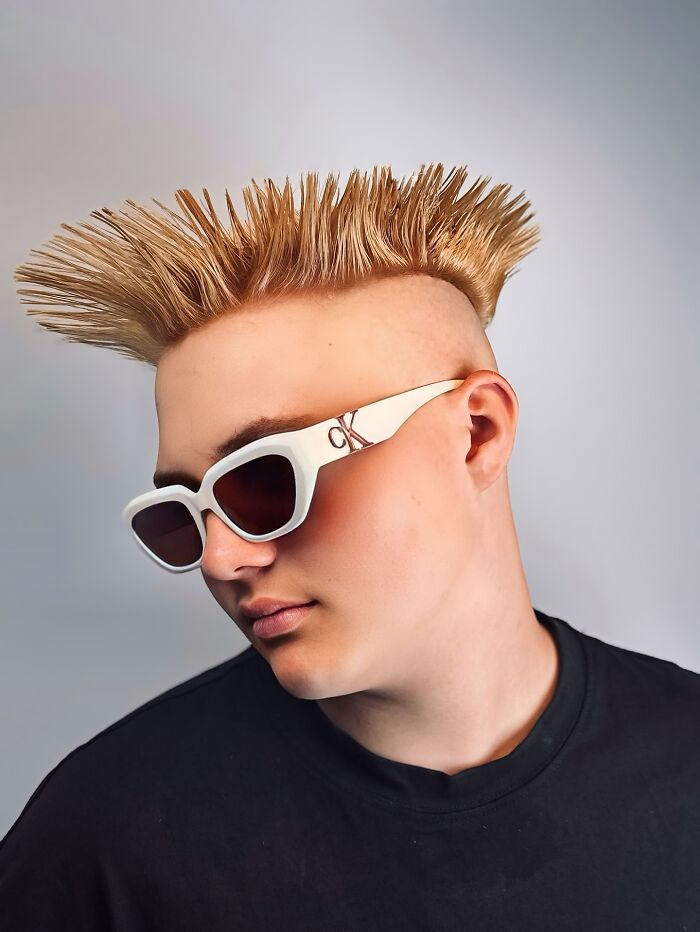 Young man with bold barber haircut styled in upright spikes wearing white sunglasses and a black shirt on a gray background