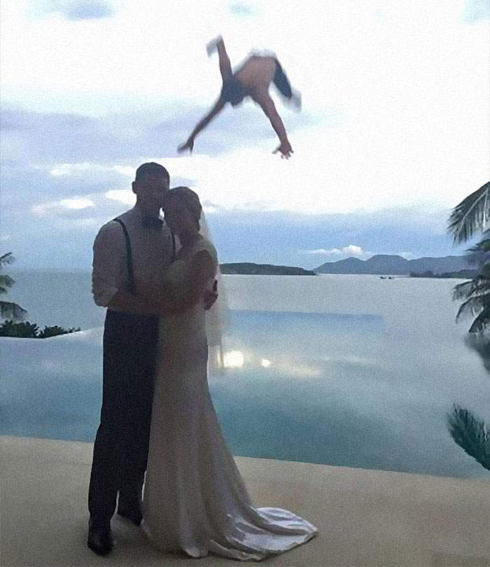 Couple embracing at a wedding near a pool as a person unexpectedly jumps in midair, captured in a frozen frame.