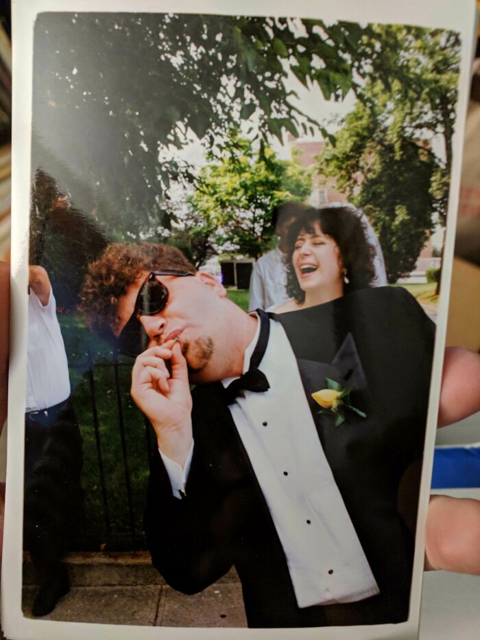 Man in tuxedo wearing sunglasses photobombs a bride laughing outdoors at a wedding with trees in the background.
