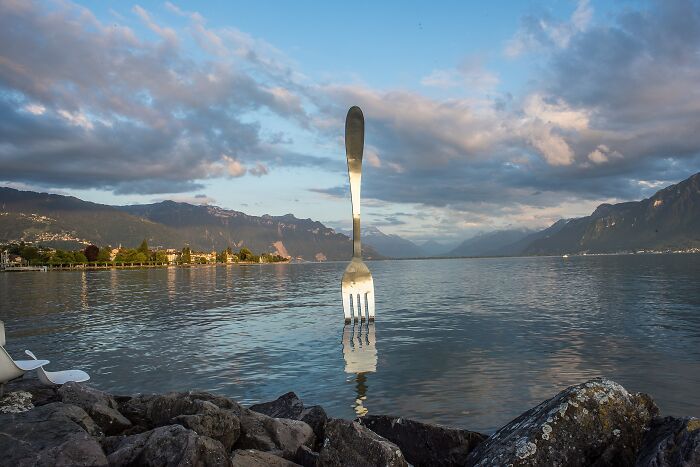 Giant fork sculpture standing in a lake with mountains and cloudy sky, a spectacular sculpture for travel bucket list.