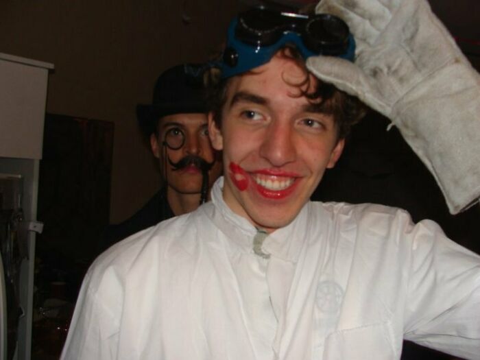 Young man in goggles and white gloves with lipstick marks, while another person photobombs wearing a hat and fake mustache.
