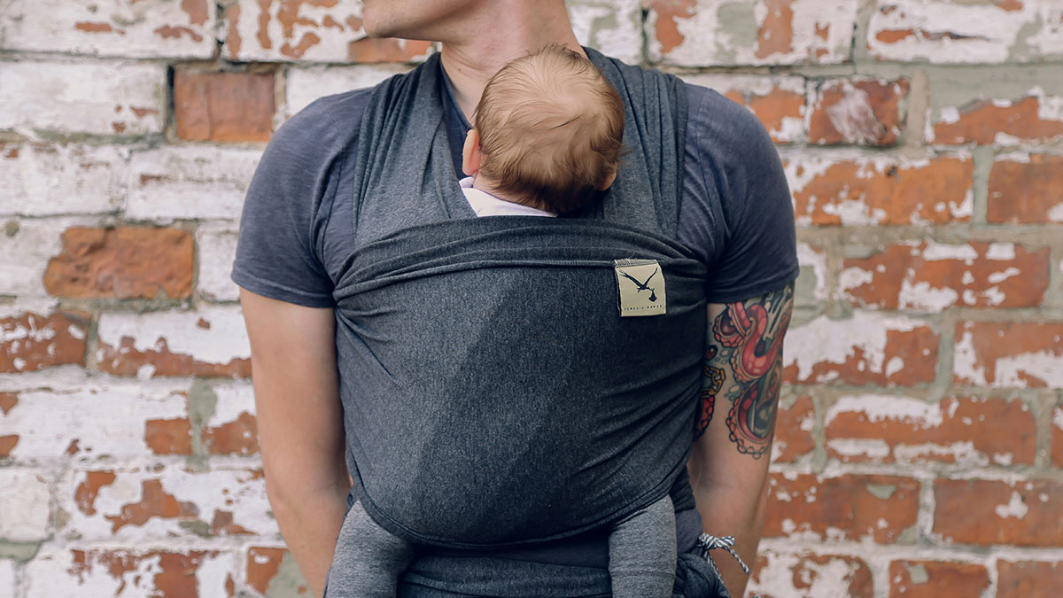 Man carrying baby in a gray wrap carrier, standing against a worn brick wall background.