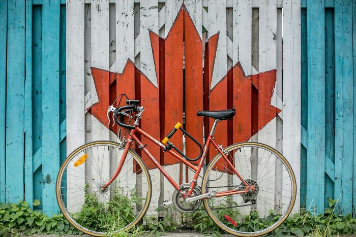 Red bicycle parked in front of a wooden fence painted with a large Canadian maple leaf, showcasing positive stereotypes.