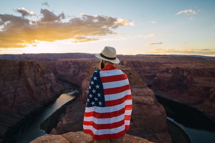 Person wrapped in USA flag overlooking canyon at sunset, showcasing great things about the US and its landscape.