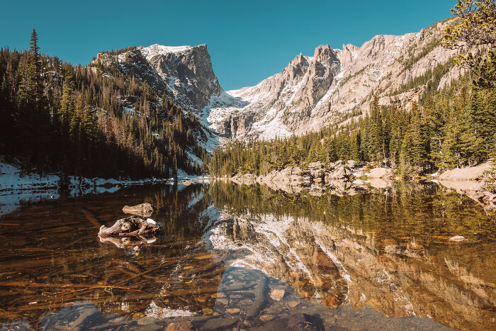 Mountain landscape in the US with clear lake reflecting snowy peaks and evergreen trees under a blue sky.