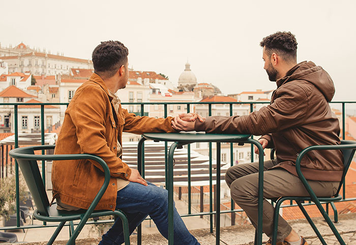 Two men sitting on a balcony holding hands and looking over city rooftops, reflecting on American expat experiences in Canada.