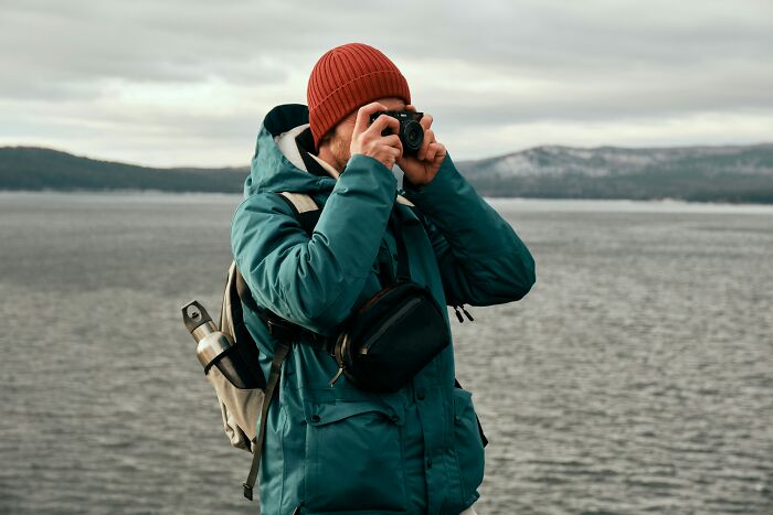 Person in a red beanie and teal jacket taking photos by a lake, symbolizing people who disappeared to start new lives.
