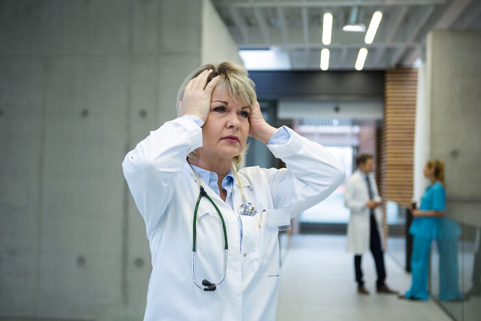 Female doctor in white coat holding head, appearing stressed while medical staff talk in hospital hallway background.