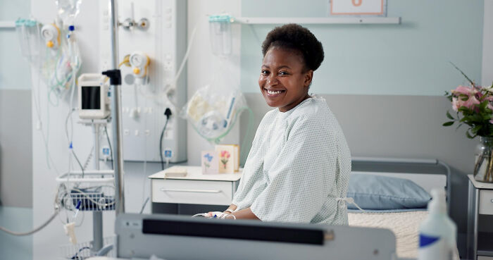 Smiling patient in hospital gown sitting on bed in medical room, highlighting anesthesia effects on patients.