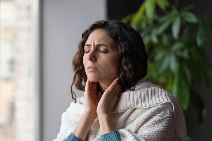 Woman with closed eyes touching neck, appearing drowsy and dazed under the effects of anesthesia in a cozy indoor setting.