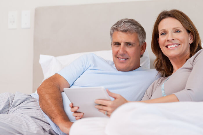 Middle-aged couple relaxing in bed looking at a tablet, illustrating patient behavior under anesthesia effects.