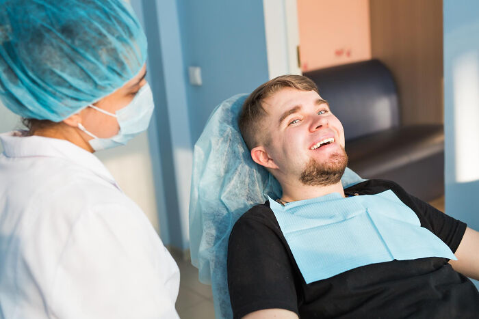 Patient smiling in a dental chair while a healthcare professional wearing a mask talks, illustrating effects of anesthesia.