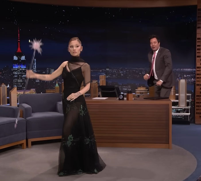 Ariana Grande in a black dress holding a wand on a late night show set during Wicked press tour.