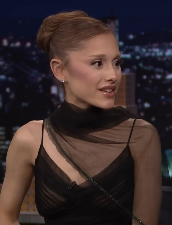 Ariana Grande in a black sheer top, speaking during a late-night show amid Wicked press tour and diagnosis reveal.