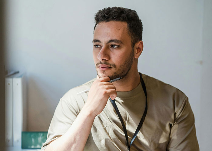 Man in a beige shirt with lanyard thoughtfully holding a pen, considering if he should ask employee about pregnancy in a right to fire state