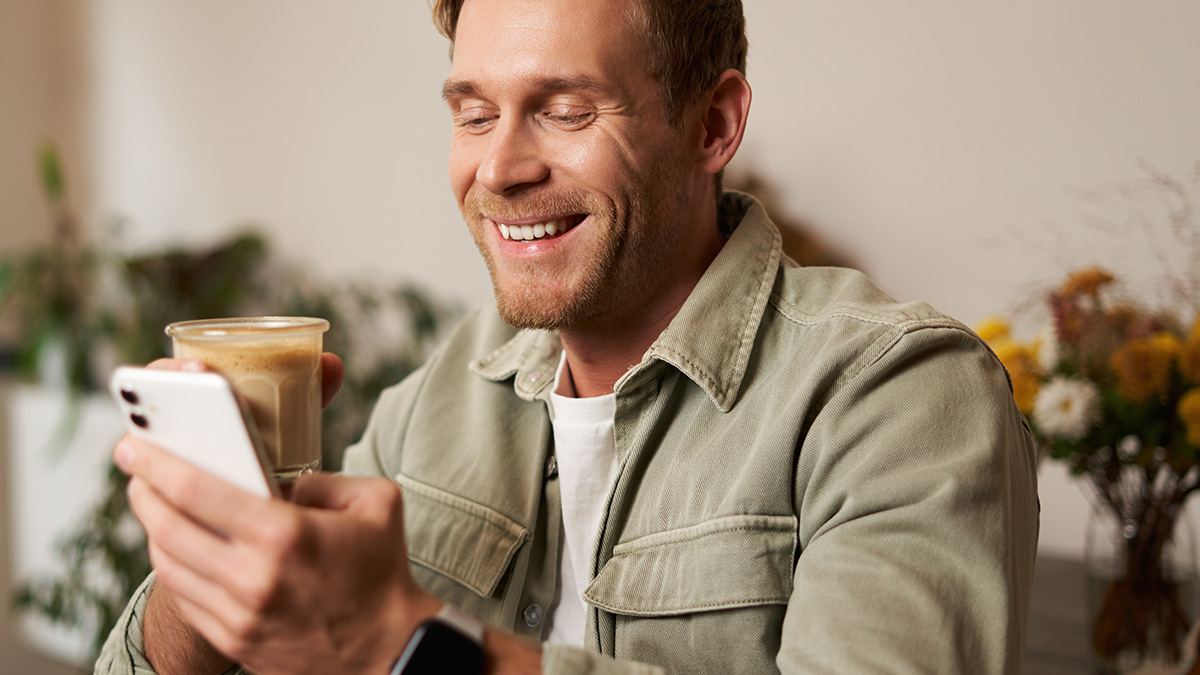 Smiling man holding coffee and looking at smartphone, enjoying funny autocorrect messages in a cozy indoor setting.