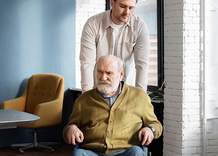 Man standing behind elderly man in wheelchair, illustrating wild family dramas and unusual relative dynamics.