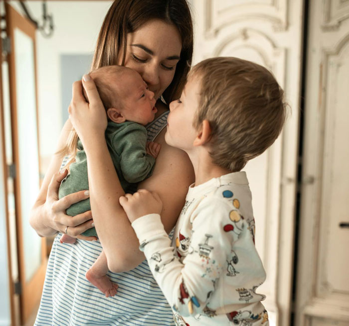 Mom holding newborn while young boy in pajamas leans in, capturing the anxiety of babysitting overnight and frequent calls.