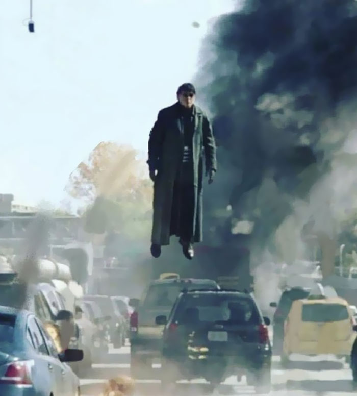 Man in long coat flying above traffic with smoke and chaos, an unhinged behind-the-scenes photo meme moment.