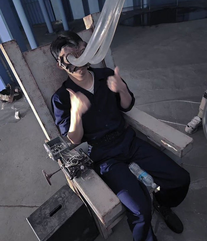 Man wearing contraption with tubes on face sitting in an unhinged behind-the-scenes setup giving thumbs up in industrial setting