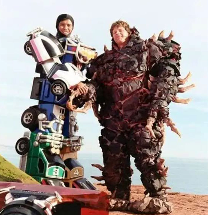 Two actors in unhinged behind-the-scenes costumes posing outdoors with a scenic ocean background.