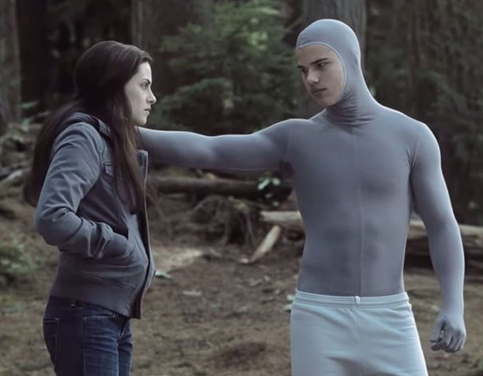 Young man in a gray bodysuit leaning against a woman in a jacket during unhinged behind-the-scenes photo shoot outdoors.